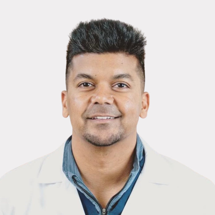 A man with short dark hair and a light beard, wearing a white lab coat over a blue collared shirt, photographed against a plain light gray background.